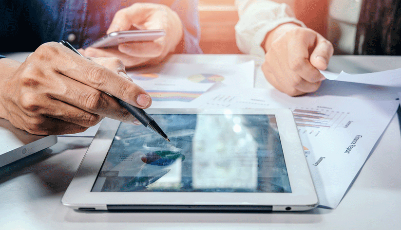closeup photograph showing hands of businessmen using a phone and a tablet while reviewing printed notes on a desk